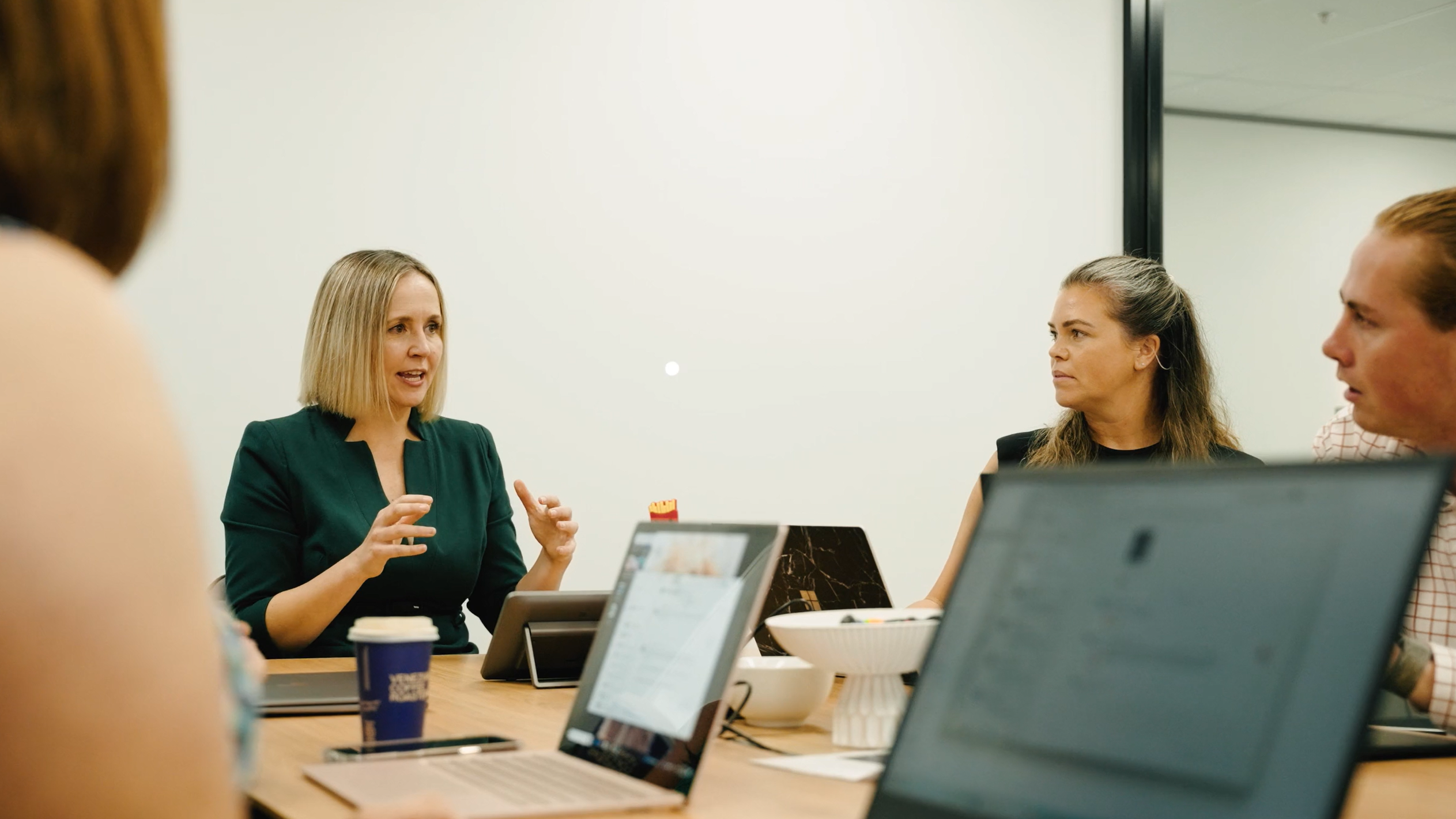A group of people in a meeting room having a discussion around a table with laptops and coffee cups.