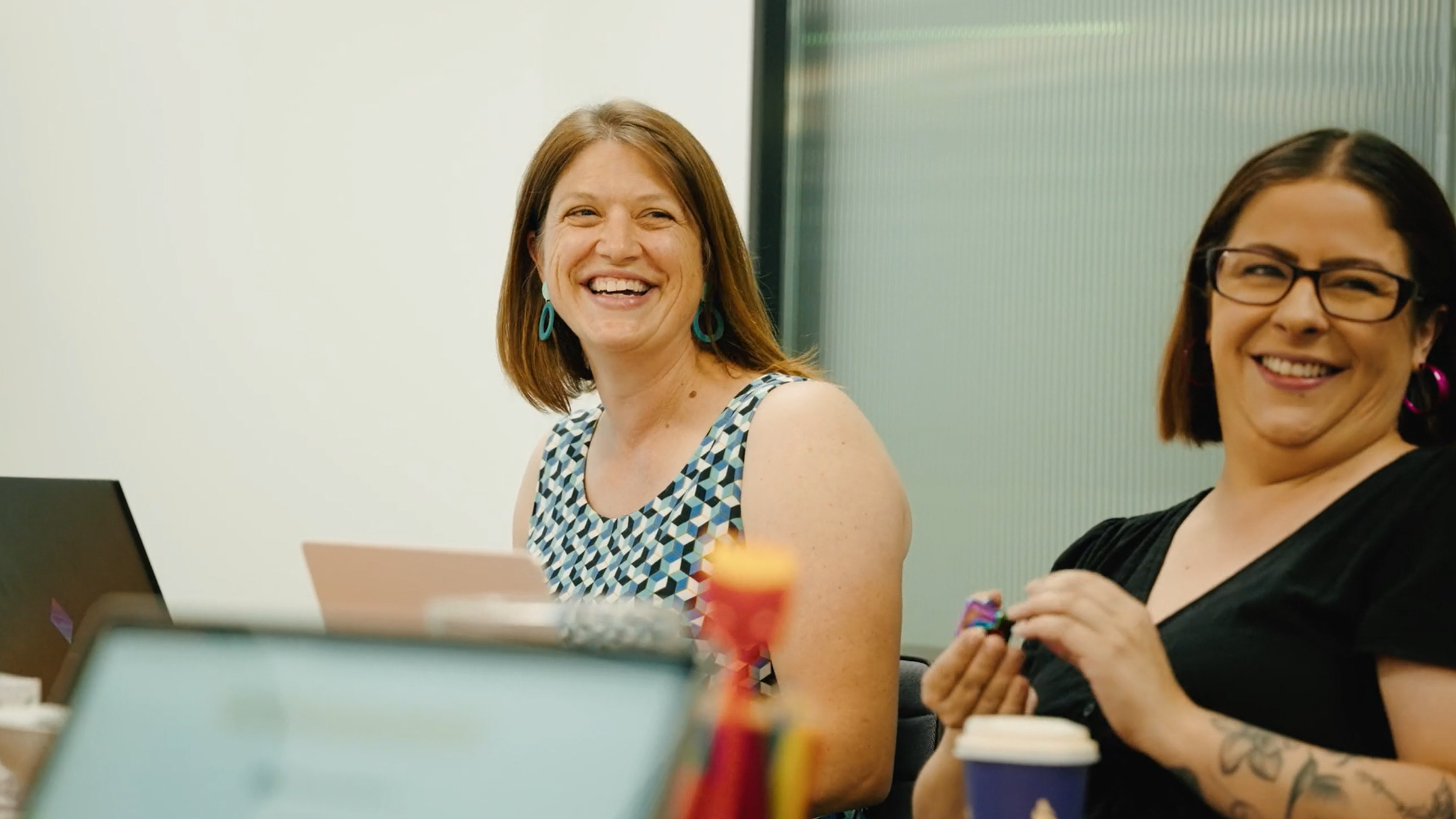 Two people sitting at a meeting table, smiling and laughing, with laptops and a coffee cup in front of them.